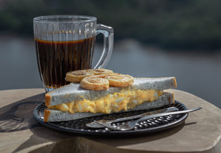 White bean butterfly shaped breads put on Butterfly pea homemade Sandwich stuffed with Egg salad served with Black coffee in glass cup on Wood cutting board with River view background. Breakfast, Coffee time in the morning, Space for text, Selective focus.の写真素材
