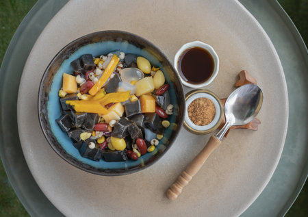 Black Grass Jelly is including Nuts, Cereals, Red beans, Ginkgo, Sliced ââpumpkin, Corn kernels, Millet, Sweet potato, Palm seed and Jelly which is served with Longan juice and Brown sugar on Ceramic tray. Traditional iced sweet dessert, Top view, Space for text,  Selective focus.の写真素材
