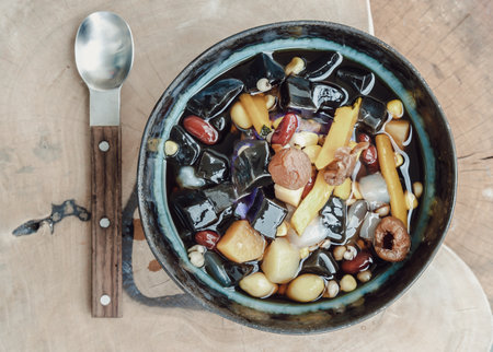 Black Grass Jelly is including Nuts, Cereals, Red beans, Ginkgo, Sliced ââpumpkin, Corn kernels, Millet, Sweet potato, Palm seed and Jelly in Longan Juice which is served with Spoon on Old wood cutting board. Traditional iced sweet dessert, Top view, Copy space, Selective focus.の写真素材