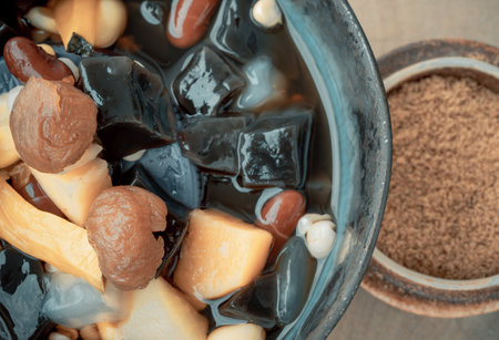 Close-up of Black Grass Jelly is including Nuts, Cereals, Red beans, Ginkgo, Sliced ââpumpkin, Corn kernels, Millet, Sweet potato, Palm seed and Jelly in Longan Juice in Ceramic bowl which is served with Brown sugar. Traditional iced sweet dessert, Selective focus.の写真素材