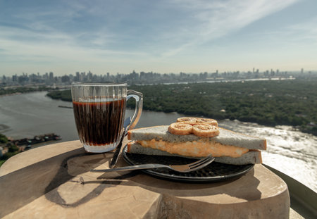 Black coffee in glass cup served with Butterfly pea homemade Sandwich stuffed with Egg salad and White bean butterfly shaped bread on Rustic wood with River view background in the morning. Breakfast, Coffee time, Space for text, Selective focus.の写真素材