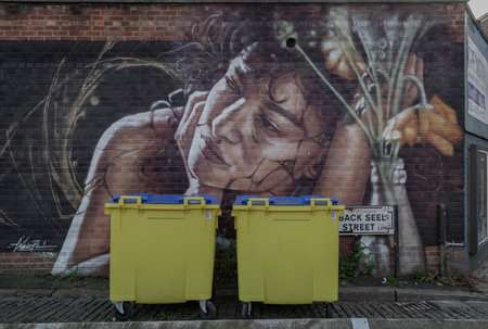 Liverpool, UK - Oct 09, 2023 - Two yellow Wheelie bin front of Portrait graffiti spray paint art...