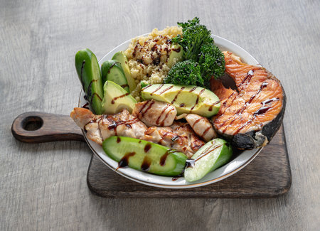 Breakfast is including Salmon steak, Chicken steak, Couscous, cut Avocado, Broccoli and cut Cucumber drizzled with Balsamic vinegar on Ceramic plate. Organic and Healthy food. Oblique view from the top, Copy space, Selective focus.の写真素材