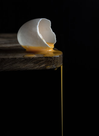A cracked raw white duck egg shell and dripping yolk on the edge of a wooden cutting board and  black background. Conceptual image of a broken white eggshell with dripping egg yolk on dark background, use it as your Wallpaper, Poster and Space for text, Selective focus.の写真素材