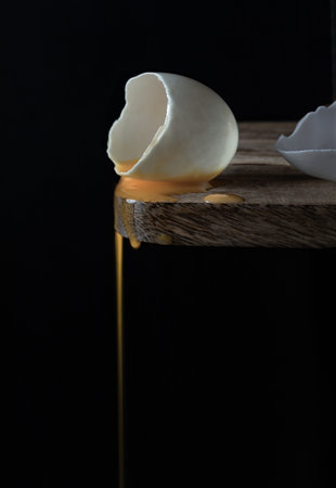 Conceptual image of a broken white eggshell with dripping egg yolk on black background. A cracked...