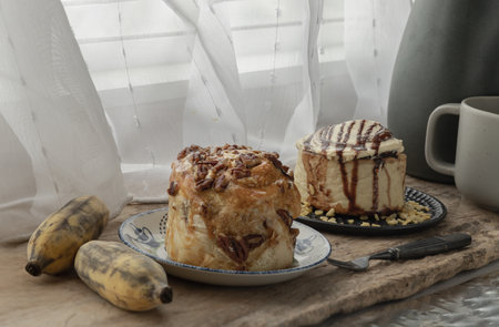 Two Homemade Caramel Pecanbon topped with caramel frosting and pecans served and Soft Chocolate Buns, Cinnamon roll topped with rich cream cheese frosting with Two Banana on old rustic wooden background by the glass window with white curtains. The concept of delicious dessert, Space for text, Selective focus.の写真素材