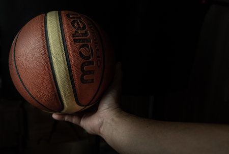 Bangkok, Thailand 14 Dec, 2024 - Silhouette view of Human hand holding basketball ball with confidence in minimalist style on black background. Close-up of A basketball leather in hand of a man against dark background, Light and Shadow, use it as your Wallpaper, Poster and Space for text, Selective focus.の写真素材