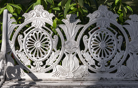 Detail of White painted cast iron victorian style garden bench in city park. Sunlight on backrest surface of Empty ornate antique cast iron bench in a shady area of the quiet park and unwind area.の写真素材