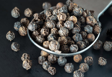 Raw organic black peppercorns or Dry black pepper seeds in stainless steel spoon on black...
