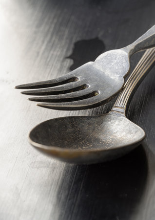 The morning sun shines hits on Ancient worn vintage cutlery on the black background. Close up of Crossed black metal spoon and fork, Highlighting its texture and design, Light and shadow, use it as your Space for text, Design, Wallpaper and Poster, Selective focus.の写真素材