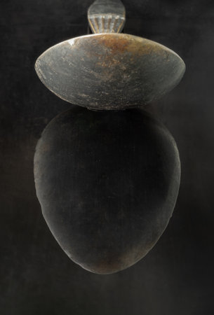 Reflection on a black surface of Old ancient worn vintage spoon in close up an oval is shaped like an egg. Curve details of Black metal vintage spoon in dark tone, Highlighting its texture and design.の写真素材