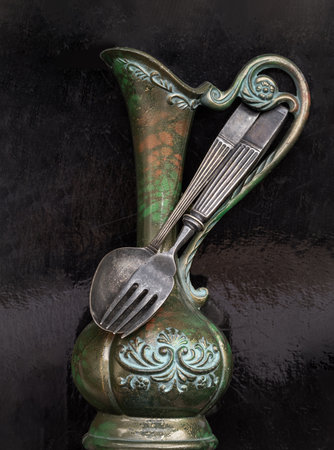 Black metal spoon and fork on Antique carved iron jug or vase with the Black background. Highlighting its texture and design, use it as your Space for text, Design, Wallpaper and Poster, Selective focus.の写真素材