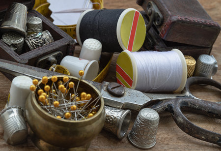 Sewing needles stuck in a white paper, Thimbles in small wooden treasure chest, Spools of black and white thread, Steel scissors and Measuring tape including pins in brass cup on wooden table. Sewing items set or Sewing objects (Sewing notions), use it as your Wallpaper, Poster and Space for text, Selective focus.の写真素材