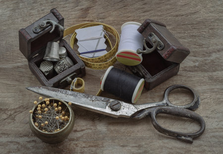 Sewing needles stuck in a white paper, Thimbles in small wooden treasure chest, Spools of black and white thread, Steel scissors and Measuring tape including pins in brass cup on wooden table. Sewing items set or Sewing objects (Sewing notions), use it as your Wallpaper, Poster and Space for text, Selective focus.の写真素材