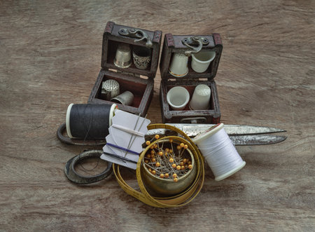 Thimbles in small wooden treasure chest, Sewing needles stuck in a white paper, Spools of black and white thread, Steel scissors and Measuring tape including pins in brass cup on wooden background. Sewing items set or Sewing notions (Sewing objects), use it as your Wallpaper, Poster and Copy space, Selective focus.の写真素材