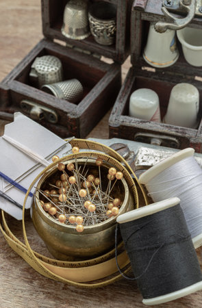 Sewing needles stuck in a white paper, Thimbles in small wooden treasure chest, Spools of black and white thread, Measuring tape and Steel scissors including pins in brass cup on wooden background. Sewing items set or Sewing objects (Sewing notions), use it as your Wallpaper, Poster and Space for text, Selective focus.の写真素材