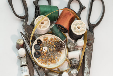 Pins and Buttons in small round tray with Steel scissors, Thimbles, Spools of thread of various colour, Measuring tape including Sewing needles stuck in a green miniature spools thread on White background. Sewing items set or Sewing notions (Sewing objects), Top view, use it as your Wallpaper, Poster and Space for text, Selective focus.の写真素材