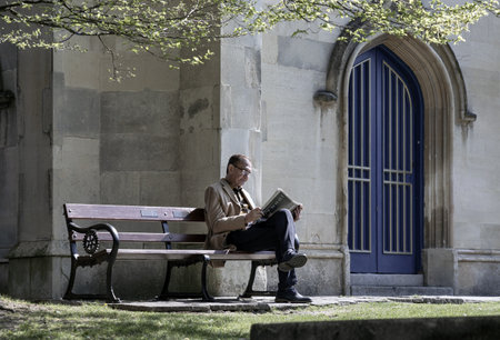 Windsor, UK - Apr 03, 2025 - An elderly man with glasses reading a newspaper while sitting on a wooden bench in the park of church. Pensioner time, use it as your Wallpaper, Poster and Space for text, Selective focus.の写真素材