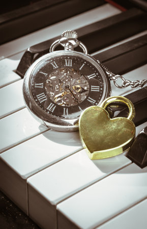 A retro pocket watch or Antique pocket watch with Golden heart shaped love padlock on Piano keys background in Vintage tone. The concept of the time of love, use it as your Wallpaper, Poster and Copy space, Selective focus.の写真素材