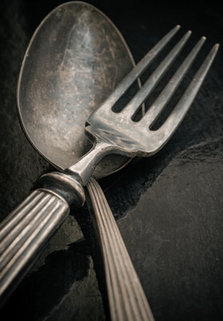 Close-up of Ancient worn vintage cutlery on the dark background. Crossed black metal spoon and fork in dark tone, Highlighting its texture and design, use it as your Space for text, Design, Wallpaper and Poster, Selective focus.の写真素材
