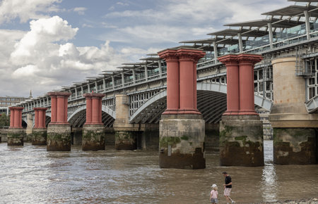 London, UK - Jul 21, 2025 - father and daughter walking along the rocks by the River Thames with red columns are remnants of an earlier railway bridge, the Blackfriars Railway Bridge and sky in the background. use it as your Wallpaper, Poster and Copy space, Selective focus.の写真素材
