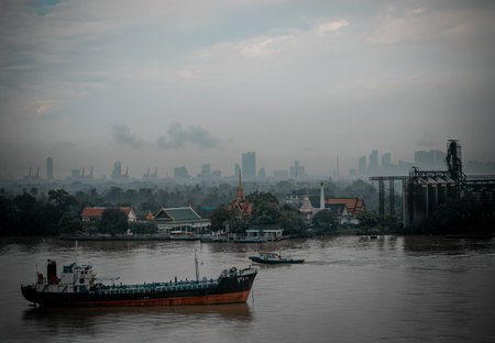 Bangkok, Thailand - 04 Oct, 2025 - a hazy atmosphere over the Chao Phraya River in Bangkok, A cargo ship and a smaller boat are visible on the water. In the background, a cityscape is visible through the haze, along with the Wat Bangkrachaonok temple and the Bang Krachao green zone. use as Wallpaper, Poster and Space for text, Selective focus.の写真素材