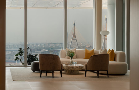Bangkok, thailand - 11 Oct, 2025 - Bathed in natural light, this elegant high-rise living space employs a neutral color scheme. Key elements include a curved off-white sofa accented with vibrant yellow pillows, two plush brown armchairs, and a sleek, rounのeditorial素材