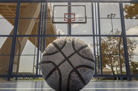 Bangkok, Thailand - 18 Dec, 2025 - A worn basketball sits on an outdoor court, the hoop and backboard visible beneath the imposing structure of the bridge. Space for text.の写真素材