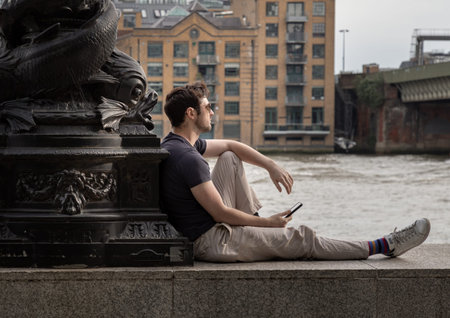 London, UK - May 31, 2025 - a moment of quiet contemplation on the banks of the River Thames, where a man rests against a weathered, ornate monument. His relaxed posture seated on a stone ledge with legs outstretched contrasts with the grand, dark metalwoのeditorial素材