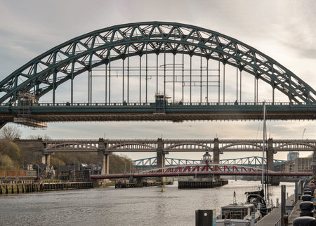 Newcastle, UK - Apr 13, 2025 - A procession of steel and stone spans the River Tyne, led by the iconic green arch of the Tyne Bridge. Its sweep frames the High Level Bridge and the red lattice of the Swing Bridge resting below. Along the quayside, moored のeditorial素材