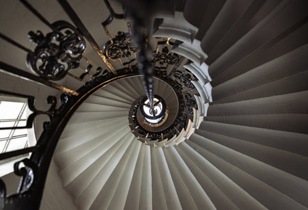 London, UK - Jun 18, 2025 - This perspective of the spiral stairs within the Chapel of St Peter and St Paul at the Old Royal Naval College, Greenwich, transforms a functional element into a stunning geometric masterpiece. Looking directly upward, the rhythmic, overlapping curves of the white stone steps create a Fibonacci-like spiral that suggests a sense of infinite ascent toward the chapel's upper levels and dome. The intricate, dark wrought-iron balustrade provides a sharp, elegant contrast to the pale masonry, while a central lantern chain anchors the composition, drawing the eye toward the light above. Balancing grace with structural precision, this staircase stands as a testament to the sophisticated architectural detail found throughout this historic site.