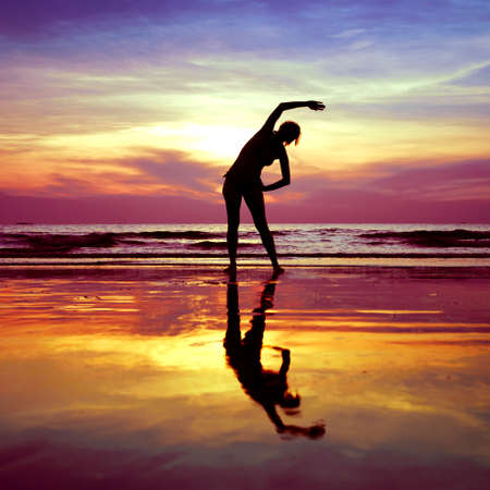 Silhouette of young woman, yoga stretching exercises on the beach at sunsetの写真素材