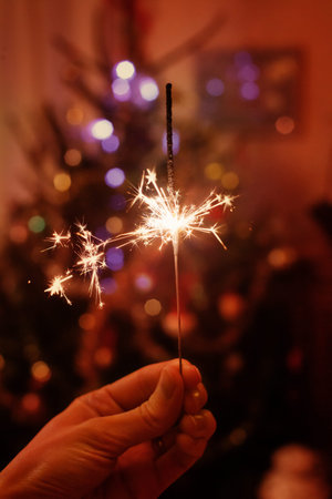 hand holding sparkler near christmas tree, new year celebration at home