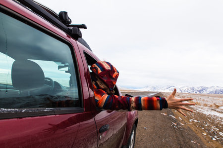 backview of girl in bright burgundy colored jacket holding hand out of the car at empty road with...