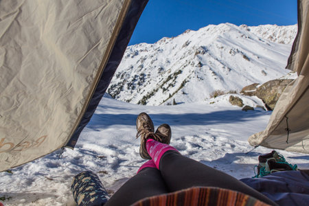 girl legs in pink socks and hiking shoes in tent with big almaty lake with snow covered hills veiw...