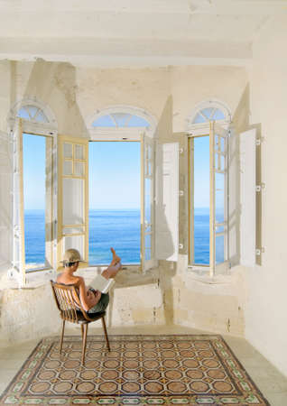 Young pregnant woman reading a book by the window facing the sea. Maltaの写真素材