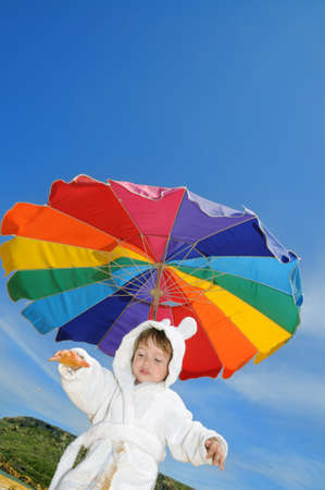 Two year old boy under colourful beach umbrella in white bathing robe tries to walk on the sand. Malta, Gozo island; copyspace on the skyの写真素材