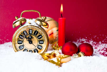 Alarm clock set at five to twelve, xmas balls under snow and a candle on dark red backgroundの写真素材