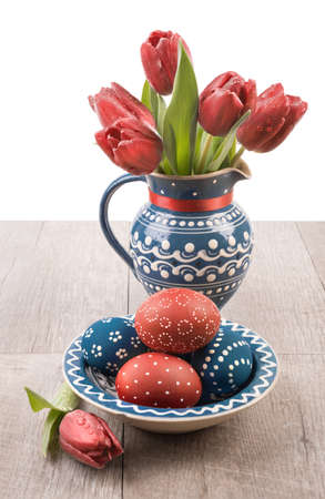 Blue ceramic vase with tulips and matching plate with painted Easter Eggs on a wooden tableの写真素材