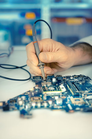 Closeup on electronic board in hardware repair shop. Shallow DOF, focus on the hand and tester tool,...
