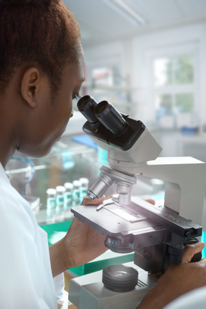 African-american female scientist or graduate student in lab coat works with a microscope in modern...