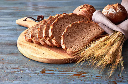 Wholegrain bread cut on board with rye buns and golden wheat ears in linen cloth on rustic wooden...
