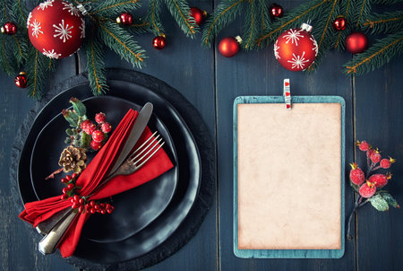 Christmas menu mockup on dark background. Black plates and vintage cutlery with Christmas decorations in green and red. Space for your text or design on white paper.の写真素材