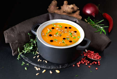 Spicy pumpkin creme soup served in ceramic pan with pumpkin oil, thyme and sunflower seeds. Dark stone background, soup ingredients around. Vegetarian dish for Autumn time.の写真素材