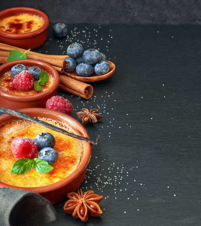 Creme brulee (cream brulee, burnt cream) with raspberry, blueberry and mint in terracota cazuela clay baking dishes on dark stone background. Border composition, space for your text.の写真素材