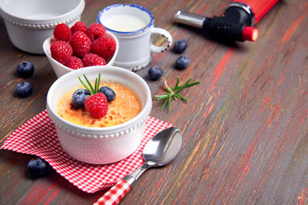 Creme brulee with raspberry, blueberry and rosemary in white ramekin with ingredients and burning torch on rustic wooden tableの写真素材