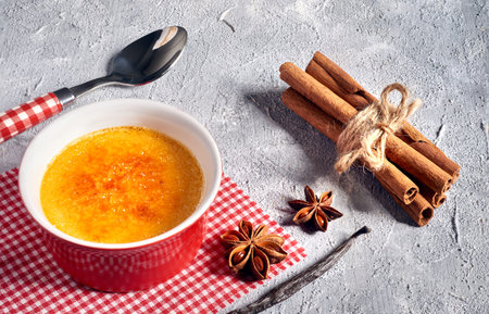 Creme brulee (cream brulee, burnt cream) on red-white checkered napkin with matching spoon, cinnamon sticks and anis stars on gray concrete backgroundの写真素材