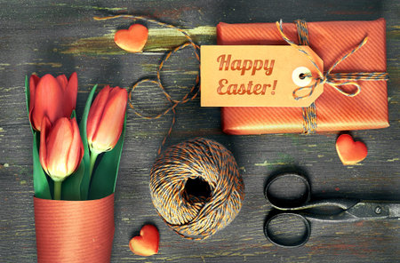Wrapped gift with tag Happy Easter, wrapping materials, scissors and red tulips on wooden table....