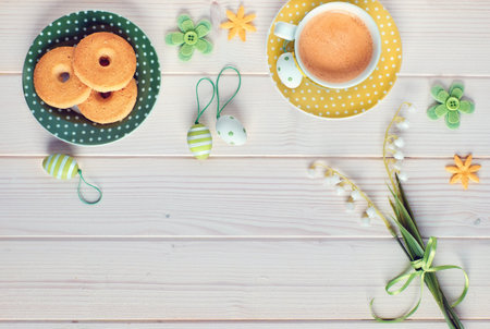 Top view on white wooden table with espresso cup, plate of cookies, Easter eggs and spring flowers....