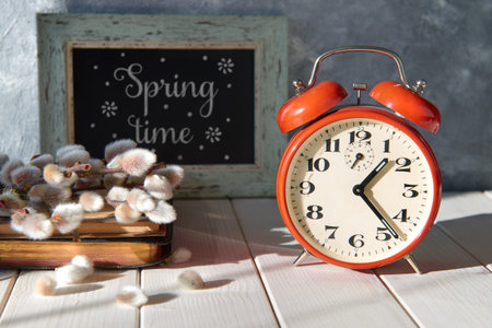 Spring sunshine. Red alarm clock, pussy-willow flowers on old book and word Springtime on...